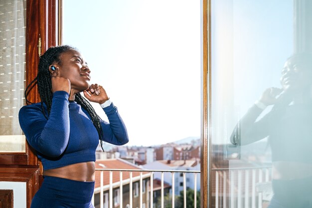 Donna afro nera vestita con abiti sportivi che ascolta musica con le cuffie in una finestra di casa sua perché inizierà ad allenarsi a casa a causa della pandemia di coronavirus covid19