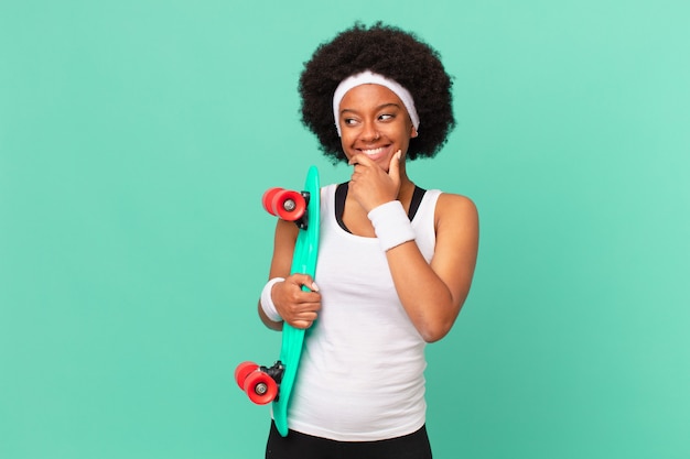 Donna afro sorridente con un'espressione felice e sicura con la mano sul mento, chiedendosi e guardando di lato. concetto di skateboard