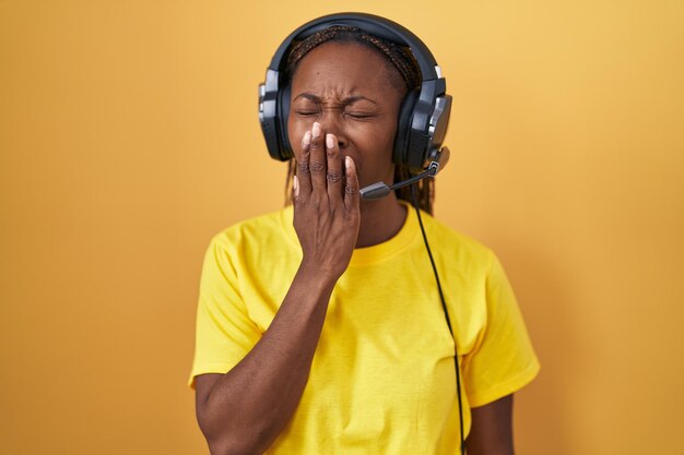 Donna afroamericana che ascolta musica usando le cuffie annoiate che sbadigliano stanche coprendo la bocca con la mano irrequieta e sonnolenza