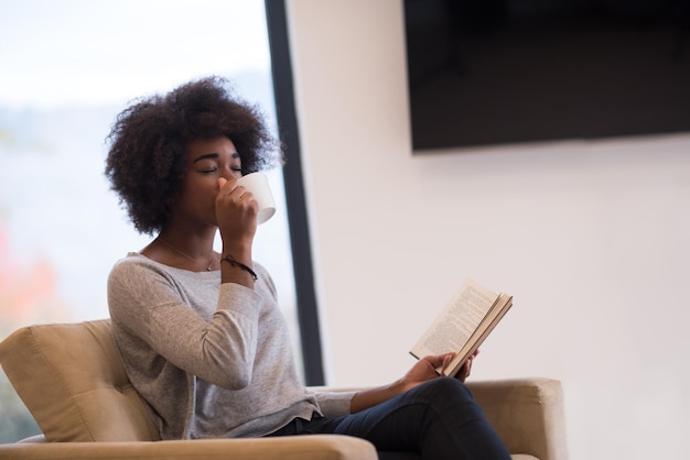 donna afroamericana che beve una tazza di caffè leggendo un libro al camino. Giovane ragazza nera con bevanda calda riscaldamento rilassante in fase di riscaldamento. autunno a casa.
