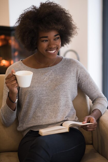 donna afroamericana che beve una tazza di caffè leggendo un libro al camino. Giovane ragazza nera con bevanda calda riscaldamento rilassante in fase di riscaldamento. autunno a casa.