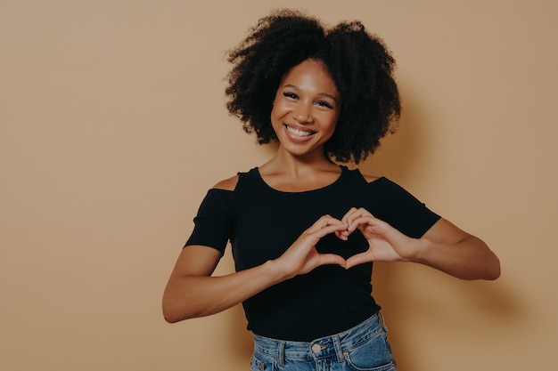 Donna afroamericana che indossa abiti casual che mostrano gesto d'amore con dita a forma di cuore, esprimendo un simbolo sano e del matrimonio con le mani. Emozioni positive e linguaggio del corpo