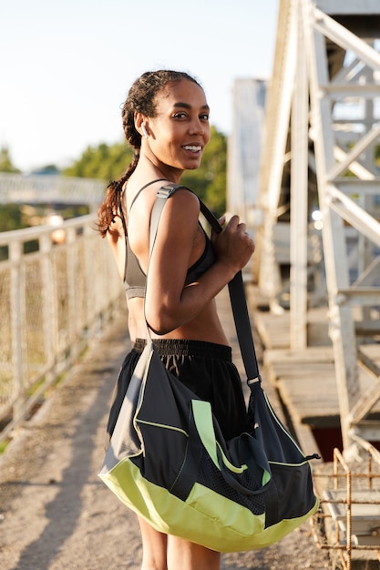 donna afroamericana che ride in abbigliamento sportivo utilizzando auricolari mentre si cammina con la borsa sul vecchio ponte