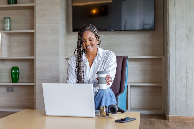 Donna afroamericana che utilizza il computer portatile in casa mentre beve il caffè seduto. Concetto di persone in casa.