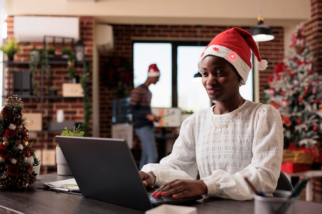 Donna afroamericana con cappello da Babbo Natale che lavora per affari in ufficio con albero di Natale e luci. Impiegato festivo che utilizza il laptop al lavoro aziendale con decorazioni e ornamenti stagionali.