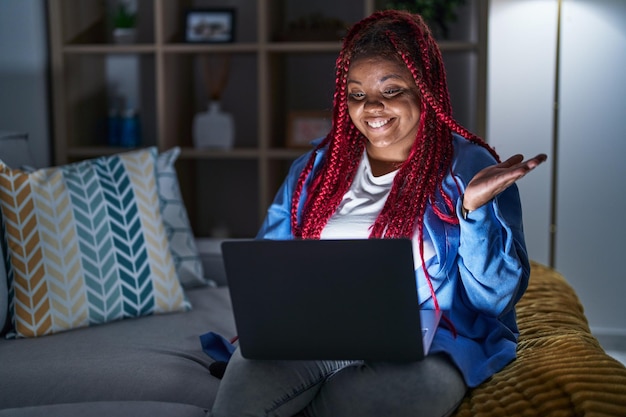 Donna afroamericana con i capelli intrecciati che utilizza il computer portatile di notte indicando da parte con le mani aperte i palmi che mostrano lo spazio della copia, presentando la pubblicità sorridendo eccitato felice