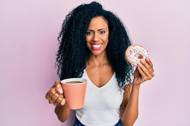 Donna afroamericana di mezza età che mangia ciambella e beve caffè sorridendo con un sorriso felice e fresco sul viso che mostra i denti