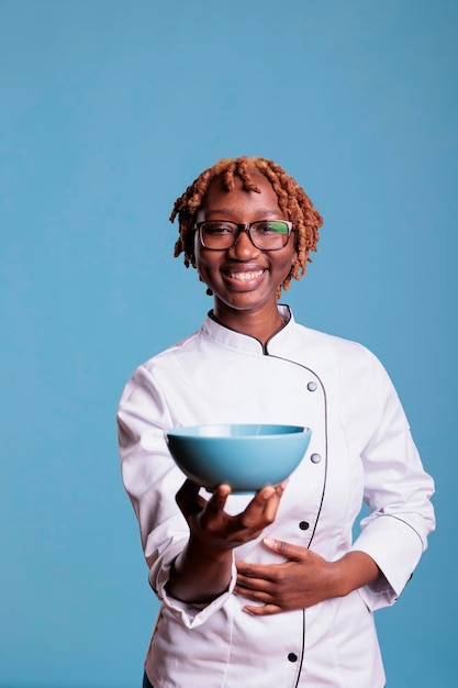 Donna afroamericana felicemente sorridente, chef professionista in uniforme con ciotola blu, ritratto in studio verticale. Fornello felice che mostra un contenitore, guardando la telecamera.