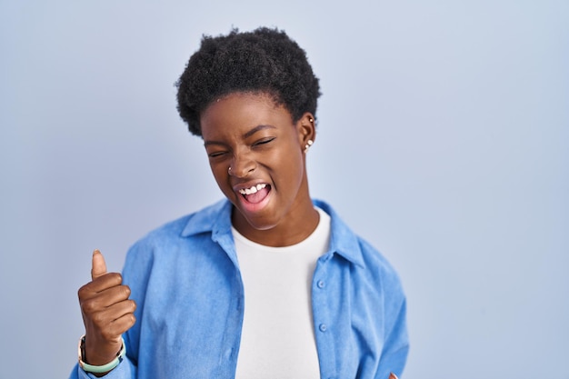 Donna afroamericana in piedi su sfondo blu molto felice ed eccitata facendo il gesto del vincitore con le braccia alzate sorridendo e urlando per il concetto di celebrazione del successo