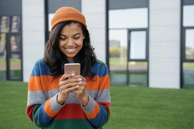 Donna afroamericana sorridente che indossa un cappello elegante utilizzando il telefono cellulare per lo shopping online per strada
