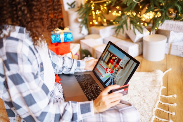 Donna afroamericana sorridente che usa il computer portatile seduto vicino all'albero di Natale con lo shopping online della carta