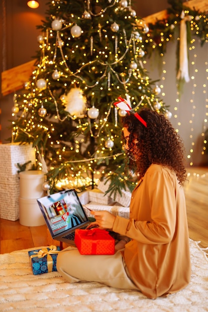 Donna afroamericana sorridente che usa il computer portatile seduto vicino all'albero di Natale con lo shopping online della carta