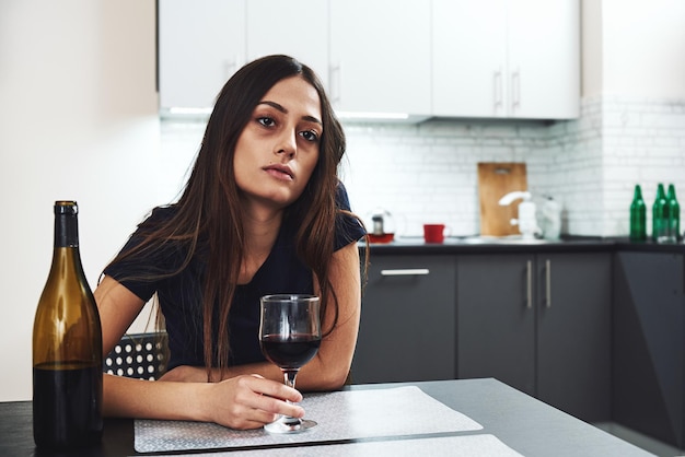 Donna alcolica dai capelli scuri, triste e sprecata seduta a casa, in cucina, bevendo vino rosso, tenendo il bicchiere, completamente ubriaca, guardando depressa, sola e sofferente postumi di una sbornia nell'alcolismo e nell'alcol