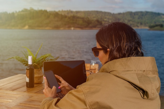 Donna all'aperto che lavora con il suo laptop e il suo telefono cellulare con un paesaggio di lago e montagne sullo sfondo
