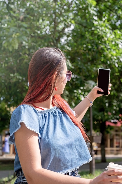 Donna alla moda allegra con capelli rossi che beve caffè al parco prendendo lo schermo del mockup del selfie