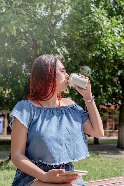 Donna alla moda allegra con capelli rossi che beve caffè al parco prendendo selfie