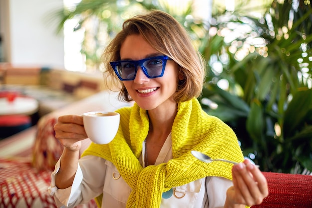 Donna alla moda che beve caffè mattutino al caffè europeo della città, accsesorizes luminosi e palme intorno.