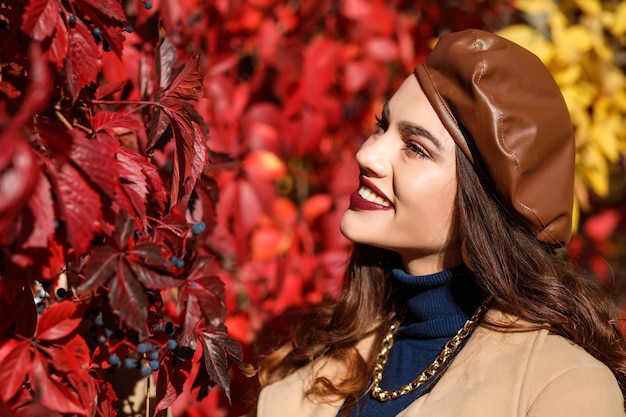 Donna alla moda che indossa un berretto in pelle marrone in piedi accanto alla siepe rossa stagione autunnale