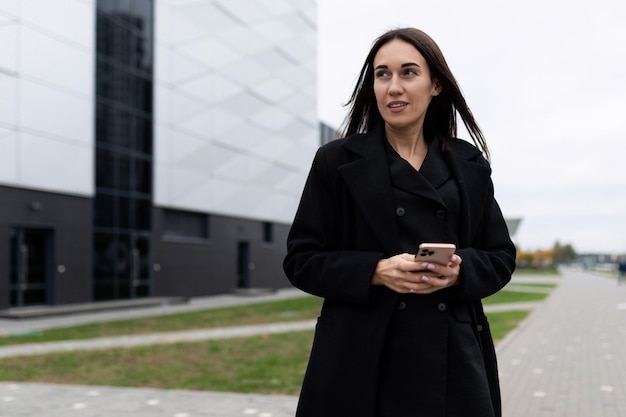 Donna alla moda di affari in un cappotto nero con un telefono cellulare sullo sfondo di un edificio moderno