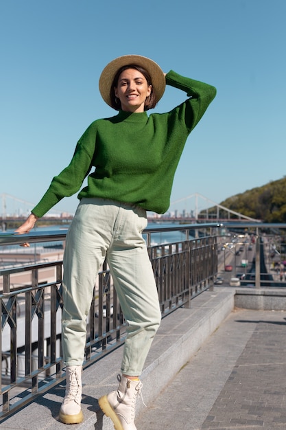 Donna alla moda in maglione casual verde e cappello all'aperto sul ponte con vista sul fiume gode di una giornata di sole estivo