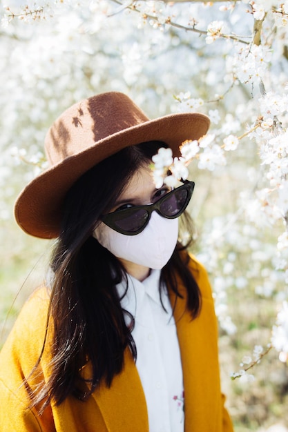 Donna alla moda in maschera fatta a mano che si rilassa da sola sui ciliegi in fiore nel soleggiato parco primaverile Elegante ragazza hipster che indossa la maschera e cammina fuori durante la quarantena