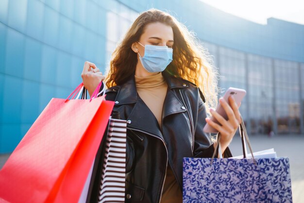 Donna alla moda in maschera medica protettiva con borse della spesa usando il suo telefono Stile alla moda