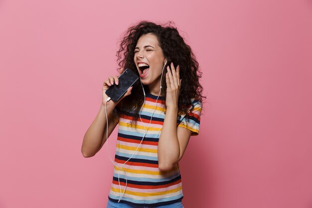 donna allegra 20s con i capelli ricci che canta mentre tiene in mano uno smartphone come un microfono e ascolta la musica tramite gli auricolari isolati su rosa