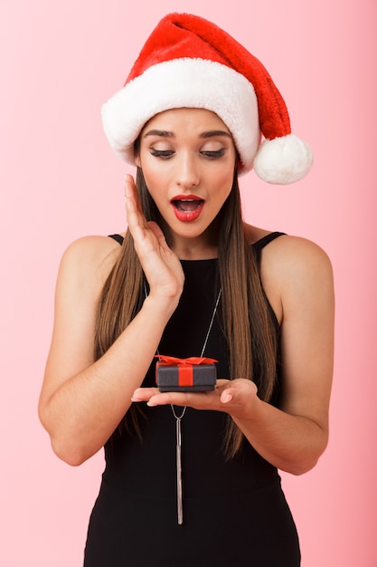 Donna allegra che porta il cappello di Natale che tiene una confezione regalo isolata su sfondo rosa