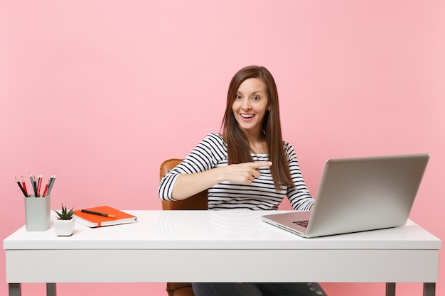 Donna allegra che punta il dito indice sul laptop pc contemporaneo mentre si lavora, sedersi alla scrivania in ufficio isolato su sfondo rosa pastello. Concetto di carriera aziendale di successo. Copia spazio per la pubblicità.