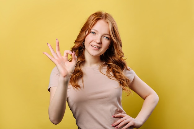 Donna allegra con capelli rossi in maglietta che sbatte le palpebre alla macchina fotografica e che mostra segno giusto, isolato