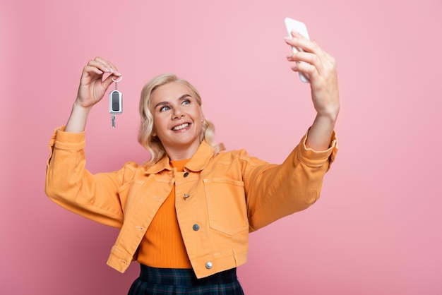 Donna allegra con la chiave dell'auto e che si fa un selfie con lo smartphone isolata in rosa