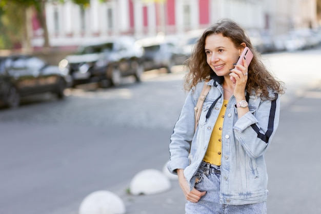 Donna allegra dei capelli ricci in giacca di jeans che parla al telefono in una luminosa giornata di sole