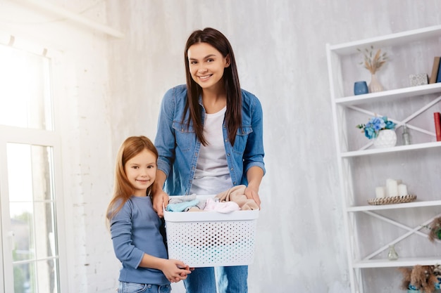 Donna allegra e sua figlia carina che tengono un cestino con i vestiti e guardano direttamente