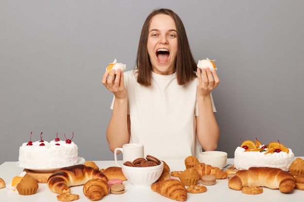 Donna allegra estremamente felice che indossa una maglietta bianca isolata su sfondo grigio seduto al tavolo festivo tra dolci che tengono torte urlando di eccitazione