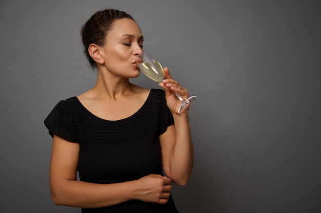 Donna allegra in abito nero beve vino spumante da un bicchiere, in piedi isolato su sfondo grigio muro con copia spazio per la pubblicità