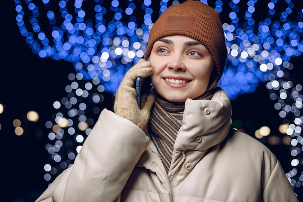 Donna allegra in capispalla invernale che parla al telefono cellulare mentre si sta in piedi contro ghirlande luminose