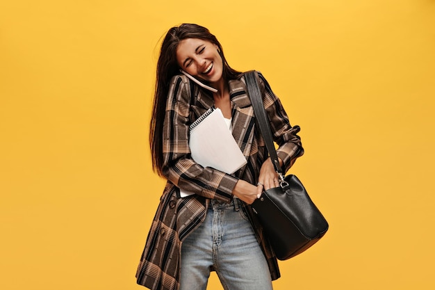 Donna allegra in jeans e giacca oversize che ride su isolato Bella ragazza parla al telefono tiene quaderni e borsa nera su sfondo giallo isolato