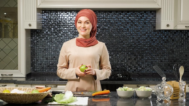 Donna allegra in uno spettacolo culinario di blog hijab che racconta agli abbonati la ricetta dell'insalata di frutta in piedi nel primo piano della cucina di casa