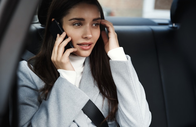 Donna allegra in vestito di affari che si siede sul sedile posteriore della sua auto e parla al telefono cellulare