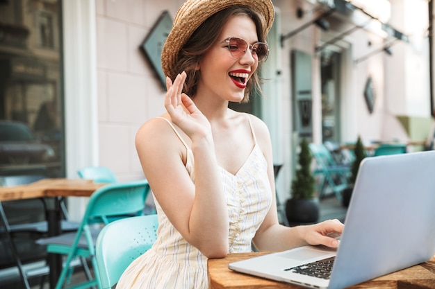 Donna allegra in vestito e cappello di paglia utilizzando il computer portatile