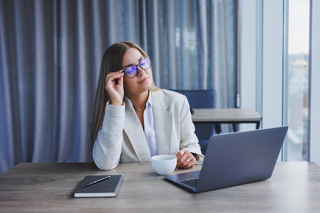 Donna allegra manager con gli occhiali con il computer portatile che sorride alla macchina fotografica durante la pausa caffè nell'interno dell'ufficio donna europea felice con bevanda alla caffeina in posa