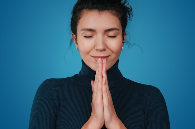 Donna allegra positiva che tiene le mani nel gesto di preghiera che si posa su sfondo blu La donna crede in una fede migliore credenza Primo piano ritratto