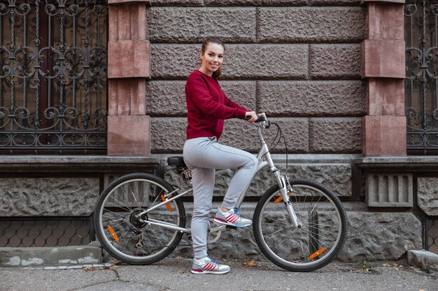 Donna allegra vestita con un maglione che cammina con la sua bicicletta in città mentre si trova vicino al muro e sorride