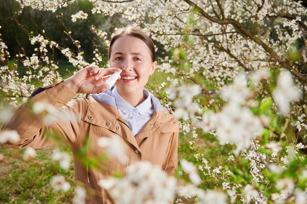 Donna allergica che soffre di allergia stagionale in primavera sorridente in giardino fiorito in primavera usando gocce nasali tra alberi in fiore Concetto di allergia primaverile