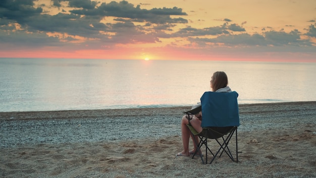 Donna ammirando il tramonto sul mare che si siede nella sedia turistica pieghevole