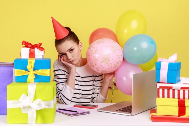 Donna annoiata che festeggia il compleanno seduta sul posto di lavoro circondata da molti regali e mongolfiere