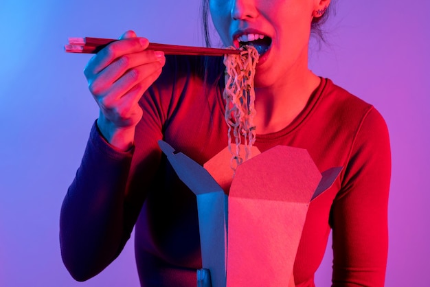 Donna anonima che tiene la scatola di tagliatelle asiatiche per il pranzo con le bacchette in studio con luci blu e rosse