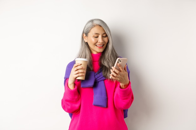 Donna anziana alla moda che beve caffè e utilizza smartphone. Signora senior coreana che legge messaggio sul telefono cellulare e sorridente, sfondo bianco.