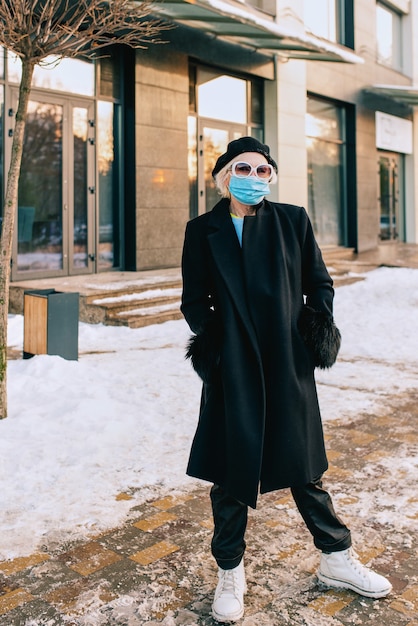 donna anziana alla moda in berretto ed elegante cappotto nero e in maschera medica che cammina all'aperto