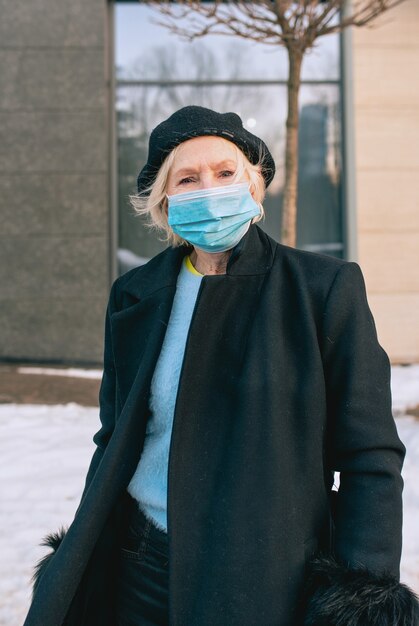 donna anziana alla moda in berretto ed elegante cappotto nero e in maschera medica che cammina all'aperto